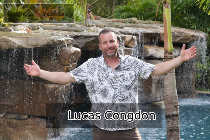 Lucas Congdon standing in front of a poolside waterfall with arms open.